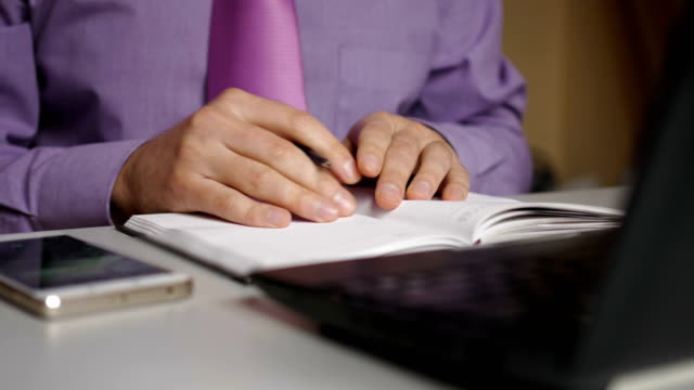 Businessman-in-a-purple-shirt-and-tie-makes-notes-in-a-notebook.-Man-writes-a-pen-in-daily.-A-person-makes-notes-in-organizer.-Laptop-and-smartphone-on-office-desk-in-office.-Closeup.
