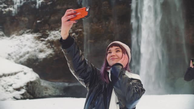 beautiful-young-woman-near-a-waterfall-in-iceland,-takes-pictures-on-the-phone,-rejoices-and-takes-a-selfie
