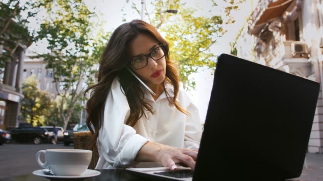 Business-female-in-glasses,-white-shirt.-Sitting-at-table-with-cup-of-coffee-and-laptop-in-roadside-cafe.-Talking-on-mobile-phone,-typing.-Slow-motion