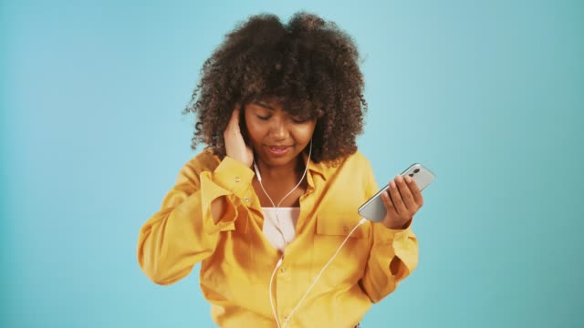 Afro-amerikanische-Dame-hören-Musik-über-Kopfhörer-mit-Smartphone-verbunden,-tanzen-und-lächelnd-posieren-auf-blauem-Hintergrund