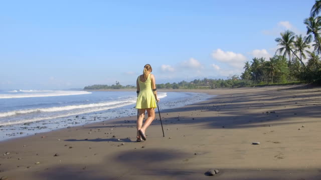 CLOSE-UP-Girl-with-crutches-walking-down-the-sandy-beach-on-tropical-island-Bali