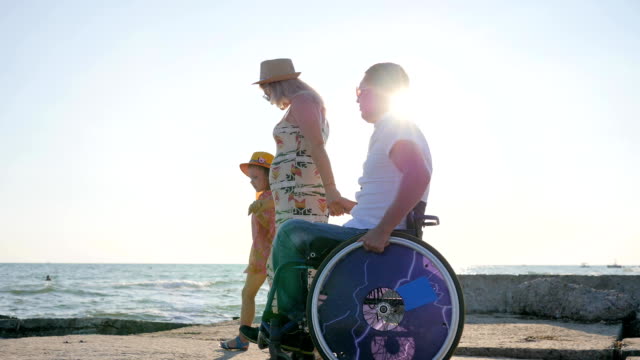 happy-disabled-Daddy-walking-with-together-with-family-beside-sea-in-backlight,-kid-with-colorful-air-balloons-walks