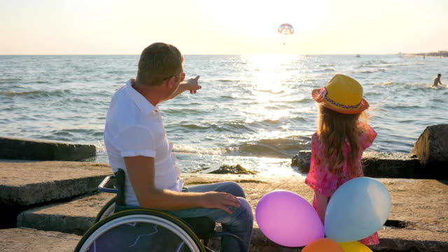 father-in-wheelchair-with-child-near-sea,-kid-hold-multi-colored-balloons