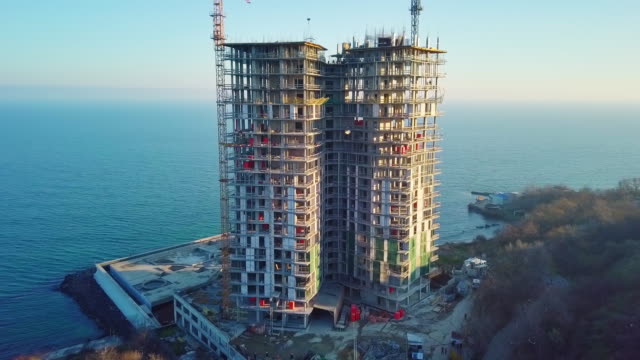 Aerial-city-view.-Construction-of-a-high-rise-skyscraper-on-the-ocean-by-two-cranes.-The-camera-moves-counter-clockwise.-From-a-low-angle