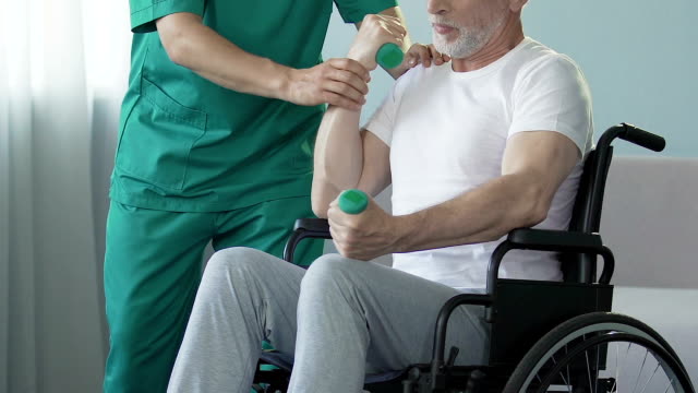 Senior-man-in-wheelchair-flexing-arms-with-dumbbells,-assisted-by-nurse,-rehab
