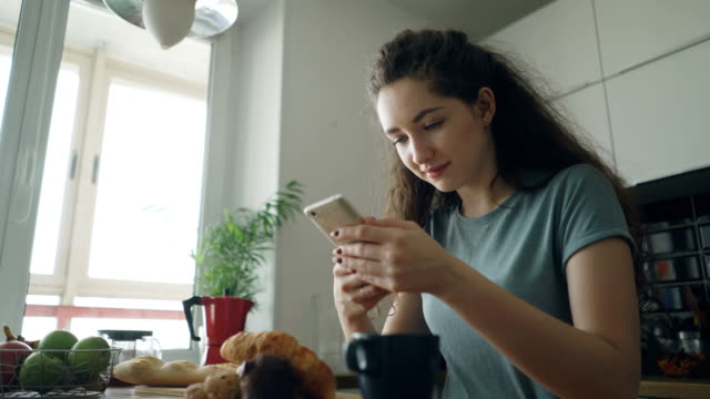 young-beautiful-curly-pretty-caucasian-woman-sitting-at-table-in-nice-kitchen-using-smartphone,-she-is-texting-someone-and-smiling,-calm-and-happy