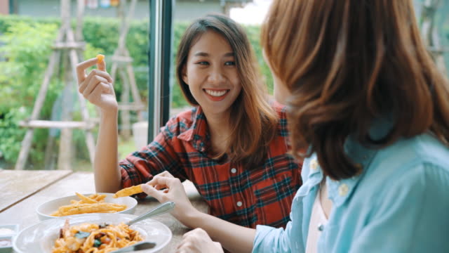 Beautiful-happy-Asian-women-lesbian-lgbt-couple-sitting-each-side-eating-a-plate-of-Italian-seafood-spaghetti-and-french-fries-at-restaurant-or-cafe-while-smiling-and-looking-at-food.