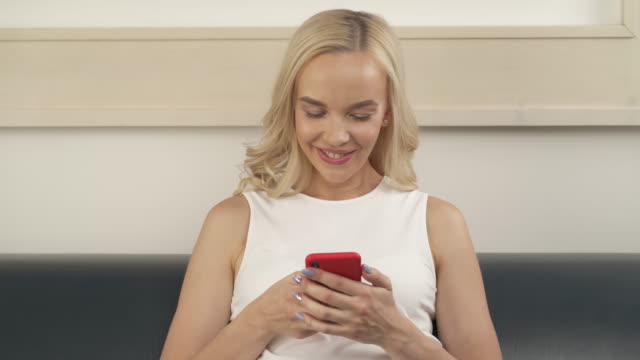 A-close-up-of-a-lovely-blonde-sitting-on-the-sofa,-looking-at-the-phone,-smiling.-The-camera-moves-from-the-left-to-the-right.