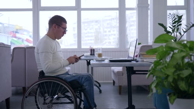 online-learning,-freelancer-man-disabled-in-wheelchair-wearing-glasses-uses-a-cell-phone-sitting-at-table-with-computer-laptop-in-a-cafe