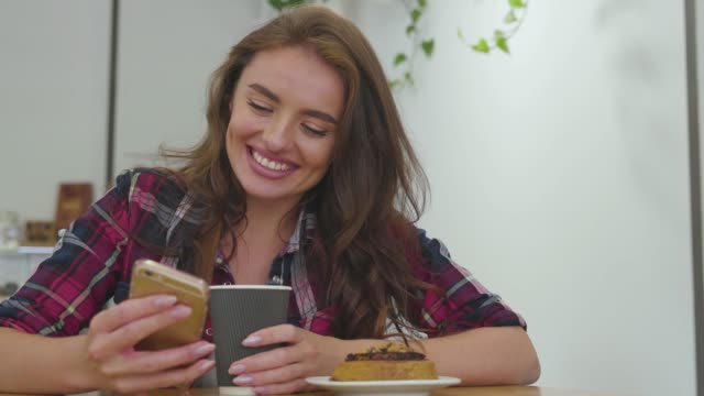 Mujer-sonriente-usando-teléfono-móvil-y-bebiendo-café-en-el-interior