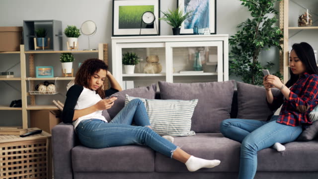 Las-amigas-en-ropa-casual-están-usando-smartphones-sentados-en-un-sofá-en-el-apartamento.-Tecnologías-modernas,-estilo-de-vida-juvenil-y-concepto-de-gadgets-de-moda.