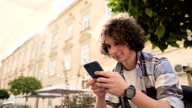 Handsome-young-man,-tourist-or-student,-millennial-in-hipster-outfit,-using-his-smartphone-scrolls-through-social-media-feed-on-device,-checking-map-or-reading-news-on-app,-looking-around,-outdoor