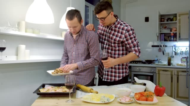Un-par-de-hombres-gays-coquetan-entre-sí-cocinando-una-pizza-juntos-y-bebiendo-un-vino.