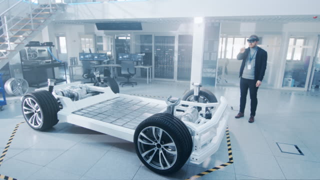 Ingeniero-automotriz-trabajando-en-la-plataforma-de-chasis-de-coche-eléctrico,-utilizando-cascos-de-realidad-aumentada.-En-el-laboratorio-de-innovación,-el-marco-del-vehículo-conceptual-incluye-ruedas,-suspensión,-motor-y-batería.
