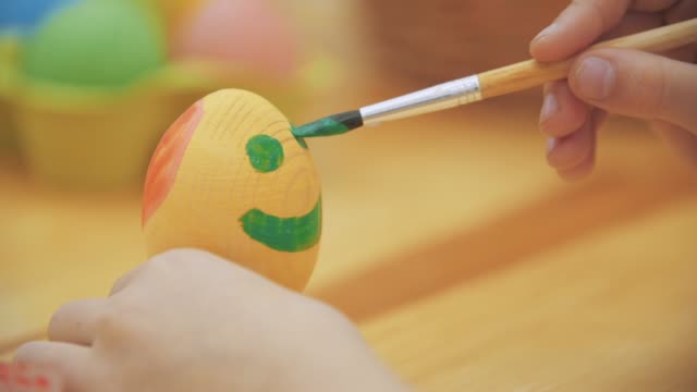 Creative-girl-chose-green-to-paint.-Lovely-girl-is-drawing-a-green-smile-on-the-Easter-egg.-Easter-concept,-table-decorated-with-a-variety-of-spring-colors.