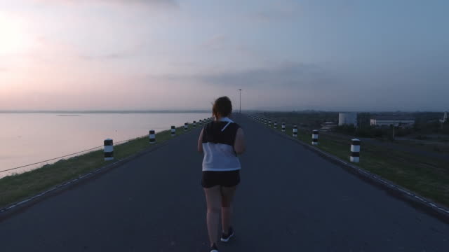 Asiatische-Frauen-joggen-in-der-Straße-am-frühen-Morgen-Sonnenlicht-am-Wasserspeicher-Pa-Sak-Jolasid-Dam.-Konzept-des-Gewichtsverlustes-mit-Bewegung-für-die-Gesundheit.-Langsame-Bewegung,-Rückansicht