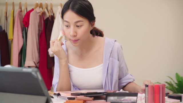 Beauty-Blogger-präsentieren-Beauty-Kosmetik-sitzen-vor-dem-Tablet-für-die-Aufnahme-von-Video.-Glücklich-schöne-junge-asiatische-Frau-verwenden-Kosmetik-Rezension-Make-up-Tutorial-übertragen-Live-Video-an-soziale-Netzwerk.