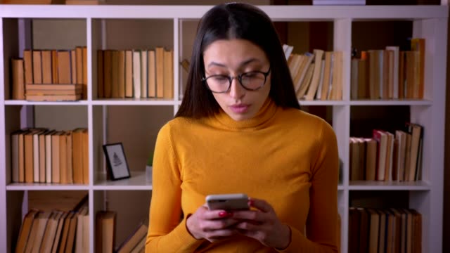 Portrait-of-beautiful-brunette-teacher-in-glasses-watching-into-smartphone-attentively-standing-at-the-library.