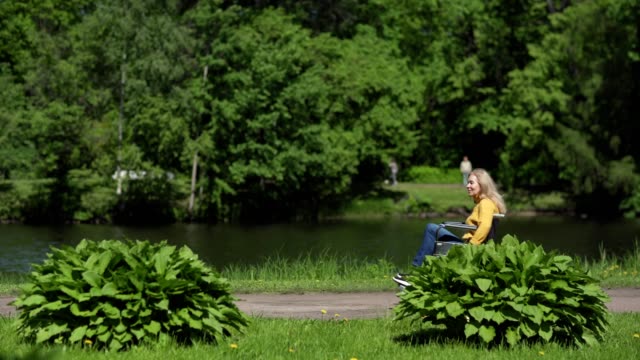 Lockdown-side-view-shot-of-beautiful-senior-woman-in-wheelchair-riding-down-path-in-green-park.-Active-disabled-female-passing-by-small-lake-on-summer-day