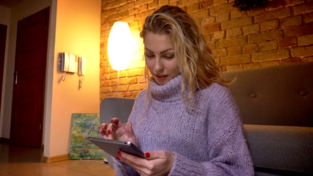 Closeup-shoot-of-adult-caucasian-blonde-female-using-the-tablet-while-sitting-on-the-floor-and-leaning-on-the-couch-indoors-in-a-cozy-apartment