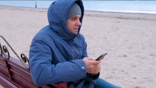 Man-blogger-in-a-blue-down-jacket-sitting-on-a-bench-on-the-sand-beach-and-writing-a-post-in-social-media-on-mobile-phone.