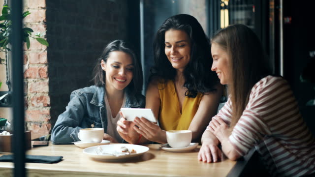 Group-of-young-women-watching-smartphone-screen-laughing-drinking-coffee-in-cafe