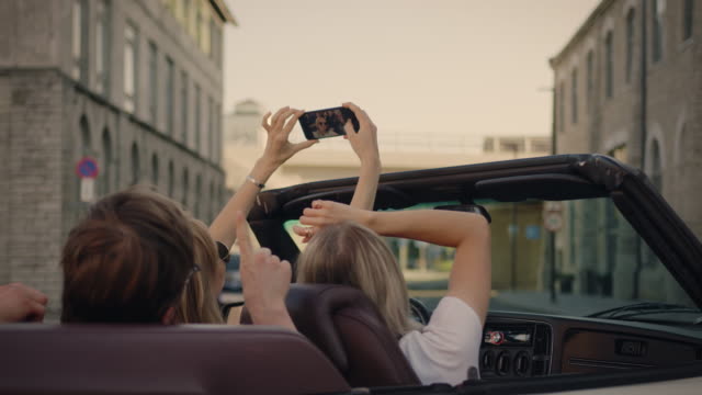 Dos-chicas-hermosas-y-dos-chicos-elegantes-enfriándose-y-divirtiéndose-en-el-coche-convertible,-usando-teléfonos-inteligentes,-tomando-selfies,-disparando-video-y-publicando-en-las-redes-sociales.-Jóvenes-Viajando,-Hablando