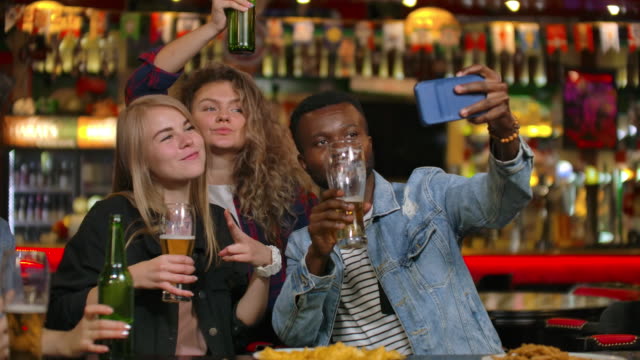 A-group-of-friends-multiethnic-resting-in-the-bar.-Friends-take-a-photo-on-the-phone-at-the-bar,-make-a-shared-photo-on-the-phone.-Party-with-friends-at-the-bar-with-beer