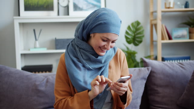 Smiling-mixed-race-girl-in-hijab-using-smartphone-touching-screen-at-home
