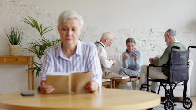 Rack-focus-of-three-elderly-people,-two-men-including-disabled-one-and-woman,-playing-cards-in-nursing-home.-Old-woman-with-short-grey-hair-reading-book-and-thinking-sadly-sitting-at-table-alone