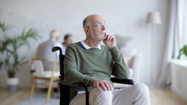 Tracking-medium-shot-of-disabled-elderly-man-in-eyeglasses-sitting-in-wheelchair-and-thinking,-then-turning-head-and-looking-sadly-at-camera-in-nursing-home