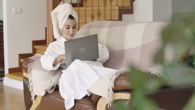 Beautiful-young-Caucasian-lady-in-bathrobe-and-hair-towel-lying-on-the-couch-and-typing-on-laptop.-Pretty-girl-using-social-media-at-home.-Cinema-4k-footage-ProRes-HQ.