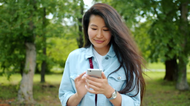 Attractive-Asian-woman-using-smartphone-in-city-park-touching-screen-smiling