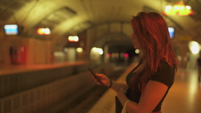 Hermosa-mujer-fresca-con-pecas,-piercings-y-pelo-rojo-mirando-el-teléfono-inteligente-en-la-estación-de-metro,-durante-el-verano-soleado-en-París.-Fondo-subterráneo-borroso.-4K-UHD.