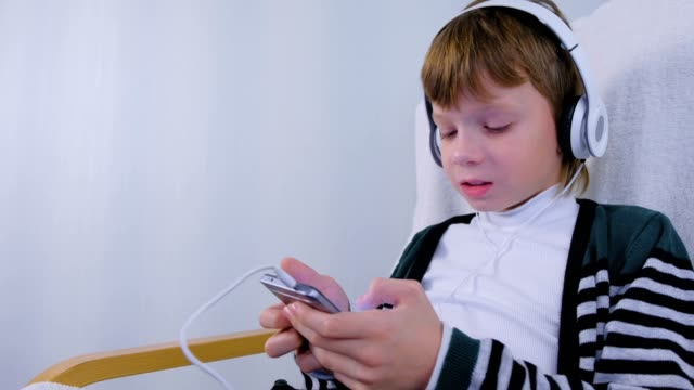 Boy-hört-Musik-in-Kopfhörern-im-Smartphone-und-singt-zu-Hause-im-Sessel-sitzend.