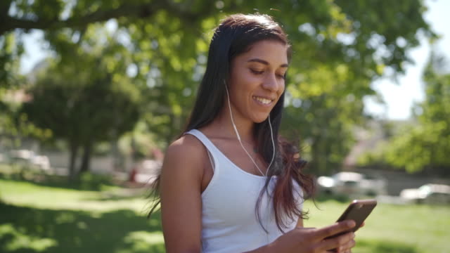Hübsche-Brünette-gemischte-Rasse-junge-Frau-hören-Musik-auf-Kopfhörer-SMS-mit-Chat-mit-ihren-Freunden-auf-Handy-im-Park