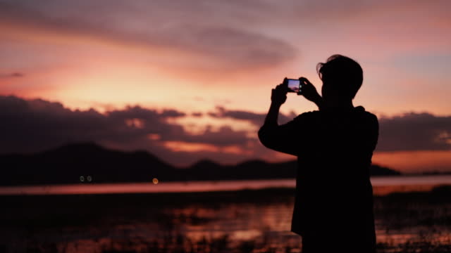 Silueta-de-retrato-asiático-hombre-usando-teléfono-inteligente-tomando-una-foto-en-la-playa-del-mar-hermosa-puesta-de-sol-de-verano,-luz-de-la-hora-dorada.