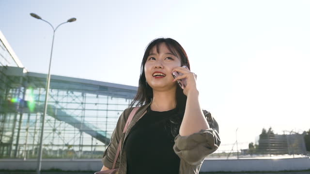 Schönes-Porträt-von-ansprechenden-jungen-asiatischen-Brünette-mit-langen-Haaren,-die-am-Telefon-in-der-Nähe-von-Flughafengebäude-sprechen
