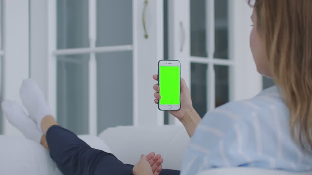 Over-shoulder-shot-of-woman-holding-a-mobile-phone-or-smartphone-with-a-vertical-green-screen-in-her-hand-at-home-lying-on-the-sofa.-Girl-tap-on-display-and-watching-content.