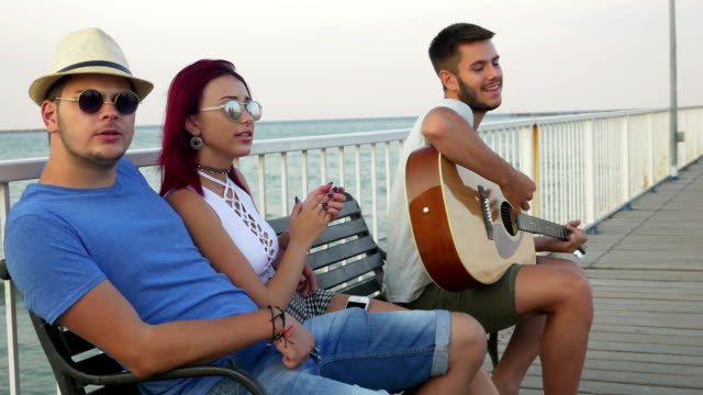 Drei-Freunde-sitzen-auf-einer-Bank-mit-dem-Meer-als-Hintergrund-und-Gesang-mit-Gitarre