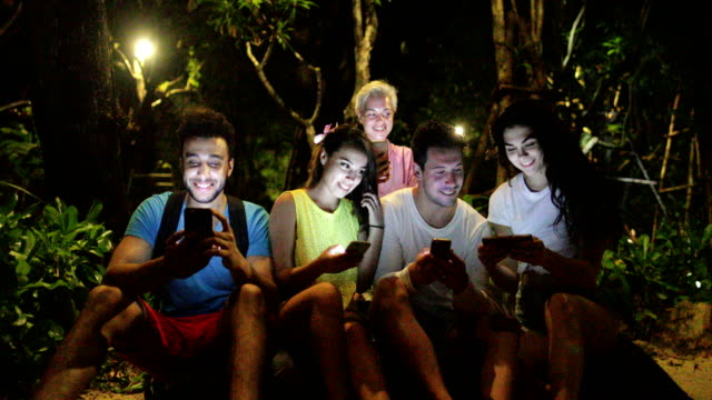 People-Group-Using-Cell-Smart-Phone-Sitting-On-Bench-In-Evening-Park,-Young-Friends-Talking-Networking-Online