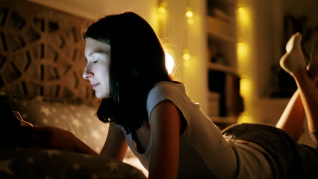 Young-smiling-woman-using-tablet-computer-for-sharing-social-media-lying-in-bed-at-home-at-night