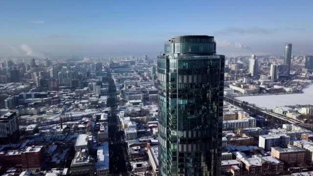 Top-view-of-the-amazing-glass-tower-or-the-business-center-in-the-background-of-a-winter-city.-Aerial-view-of-skyscraper-is-in-the-middle-of-the-city-in-winter,-blue-sky-sky-and-snowy-roofs-of-buildings-background
