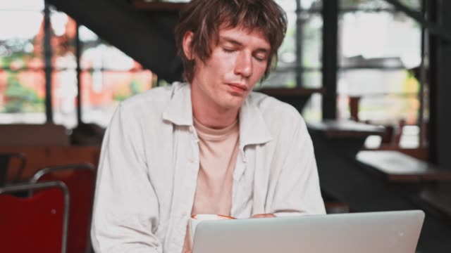 Young-attractive-cheerful-male-in-light-clothes-using-modern-portable-laptop-computer-and-drink-coffee-in-cafe.-Smiling-man-working-from-cafe-via-laptop,-social-networking-concept.-People,-communication-and-leisure.