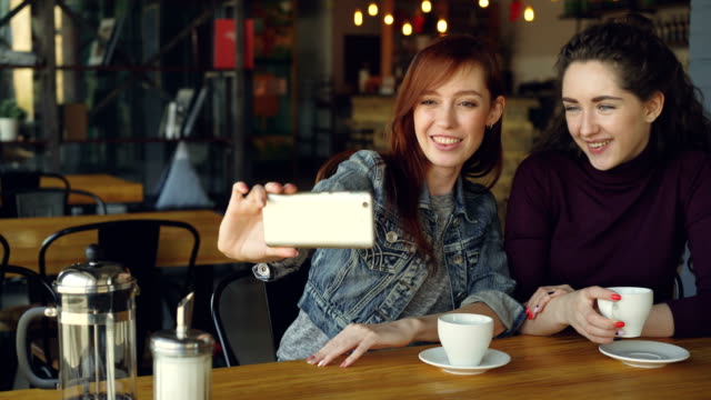 Hübsche-Mädchen-am-besten-Freunde-nehmen-Selfie-mit-Smartphone-dann-beobachten-Fotos-beim-Kaffeetrinken-im-Kaffeehaus.-Freundschaft,-social-Media-und-Spaß-Konzept.