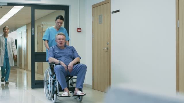 Im-Krankenhaus-Flur,-Krankenschwester-schiebt-Senior-Mann-im-Rollstuhl,-warten-Patienten-ihren-Arzt-beschäftigt-Professional-Personal-zu-Fuß.-Sauberes,-neues-Krankenhaus-mit-professioneller-medizinischer-Betreuung.