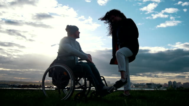 Pflege-von-Menschen-mit-Behinderungen---Mann-im-Rollstuhl-mit-Frau-sprechen-in-den-bewölkten-Sonnenuntergang
