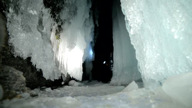 Travel-woman-on-ice-cave-of-Lake-Baikal.-Trip-to-winter-island.-Girl-backpacker-is-walking-of-ice-grot.-Traveler-looks-at-beautiful-ice-grotto.-Hiker-wears-silver-jacket,-backpack,-led-lamp.