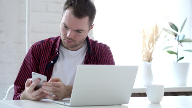 Young-Man-Using-Smartphone-while-Working-on-Laptop