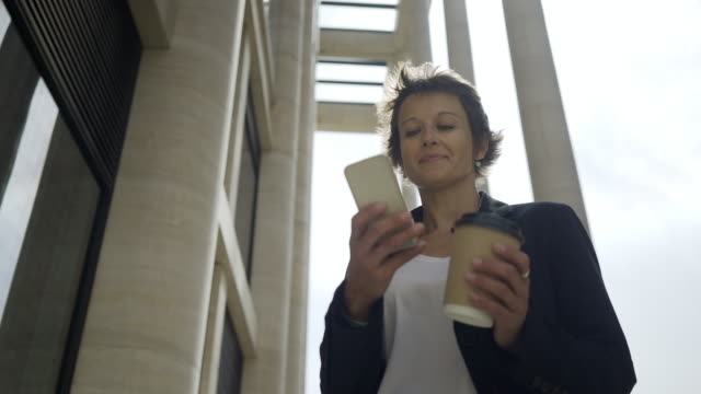 Low-angle-panning-shot-of-middle-aged-businesswoman-with-takeaway-coffee-cup-in-her-hand-text-messaging-on-smartphone-outdoors